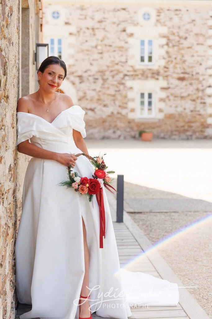 La mariée est appuyée sur le mur en pierres de la magnifique salle de réception, tout en ayant le Manoir Sainte Marie en cadre de fond. Elle est vêtue d'une magnifique robe en satin avec une légère fente laissant paraître une partie de sa jambe et ses chaussures rouges, un bouquet en forme de couronne pour rappeler son thème de Noël.