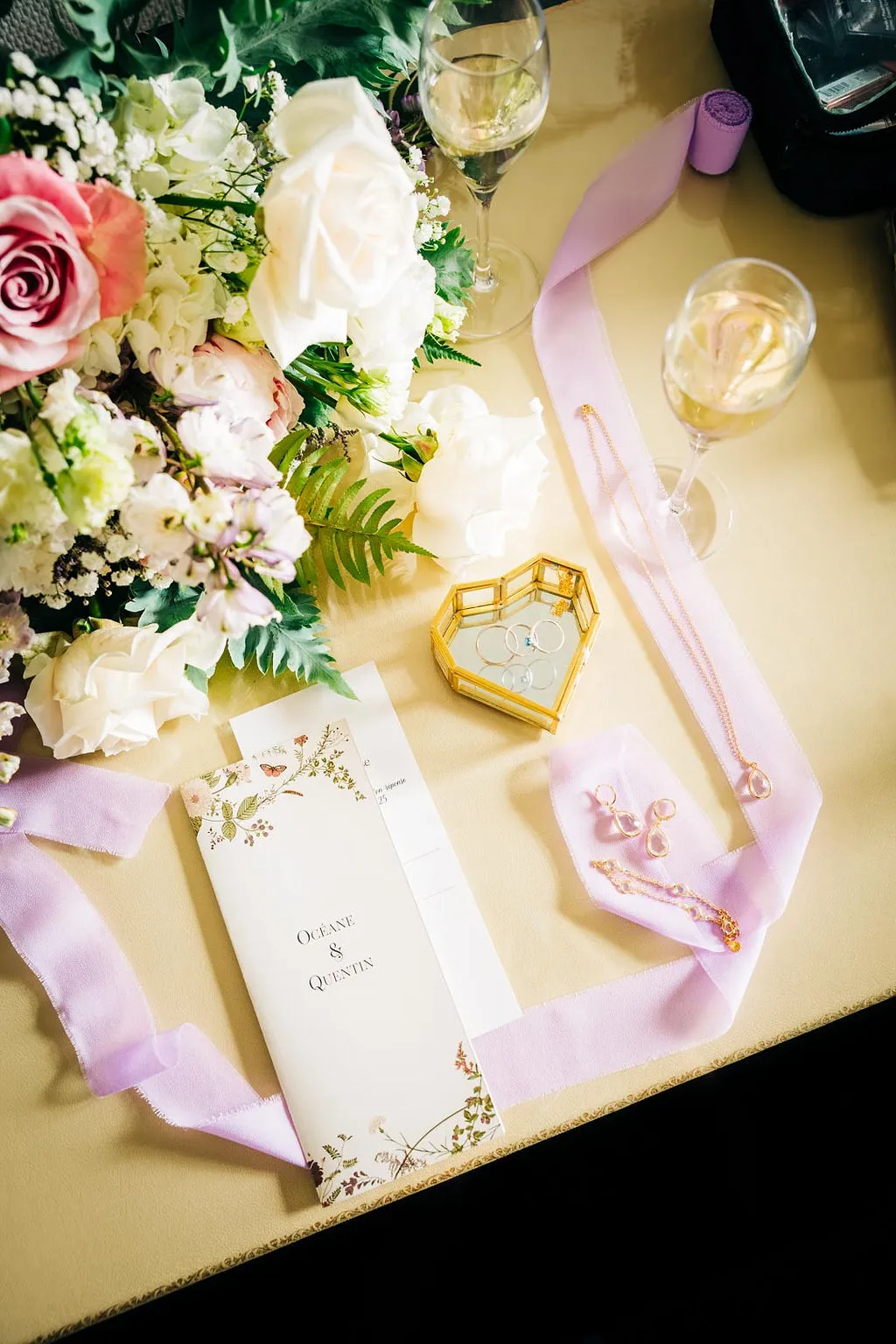 flat lay de la mariée sur le thème romantique chic,. Le faire-part est entouré de mousseline de soie parme, le bouquet destructuré de la mariée au coin en haut à gauche de l'image. La bague et l'alliance de la mariée sont posées sur un porte alliance en coeur vitré, serti de doré. Une coupe de champagne et les bijoux de la mariée sont aussi de la partie